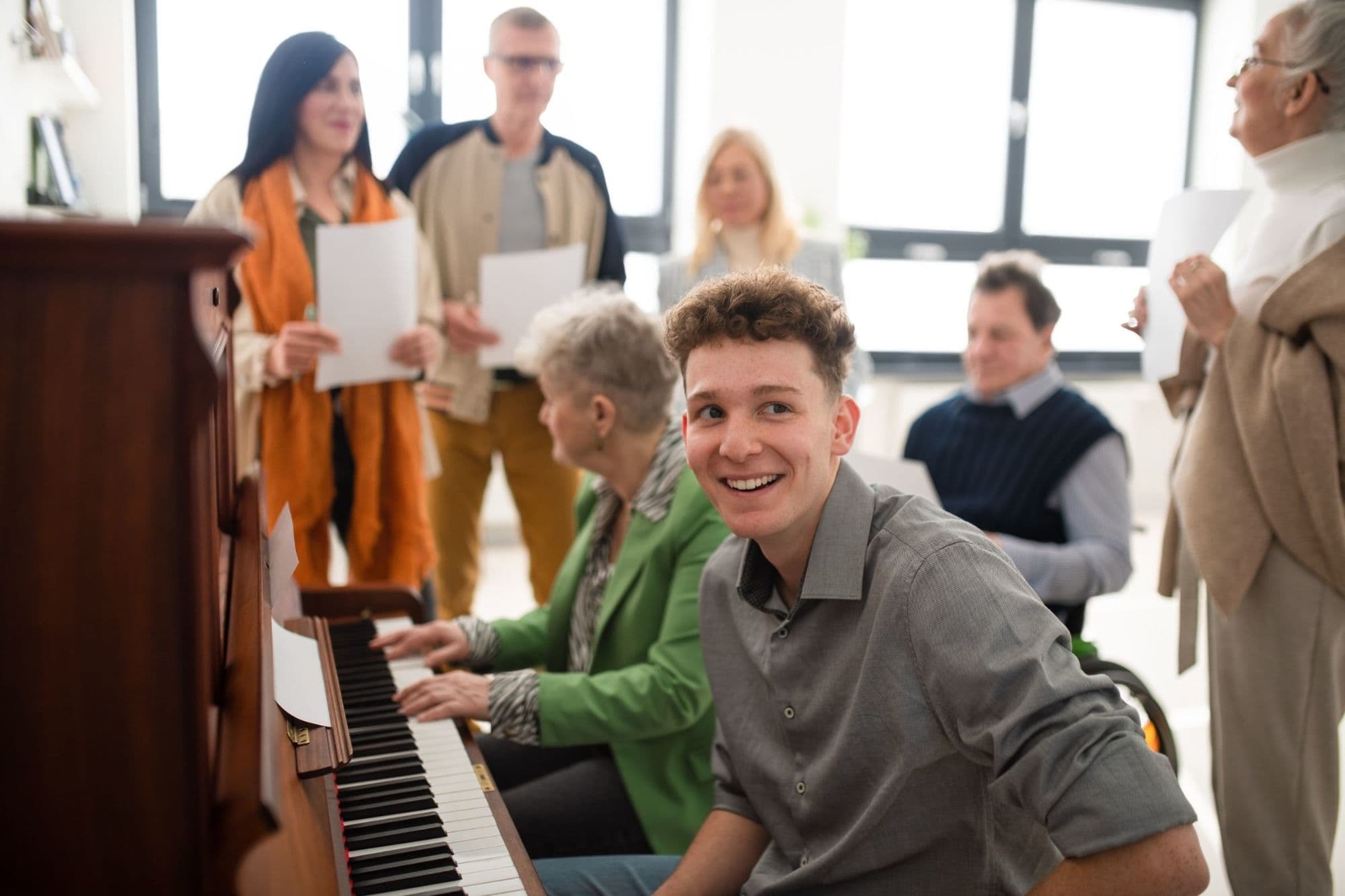 A young man at an upright piano smiles over his shoulder while an older woman plays beside him and a small group sings from song sheets.